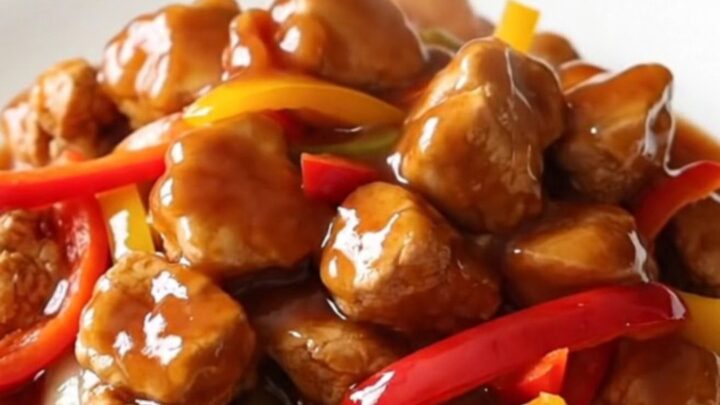 A close-up of tender Beijing chicken pieces coated in a glossy red sauce with bell peppers on a white plate.
