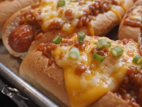 A row of golden-baked chili cheese hot dogs on a foil-lined baking sheet with bubbling melted cheese and green onions.
