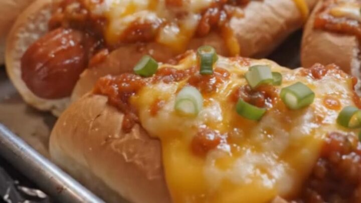 A row of golden-baked chili cheese hot dogs on a foil-lined baking sheet with bubbling melted cheese and green onions.