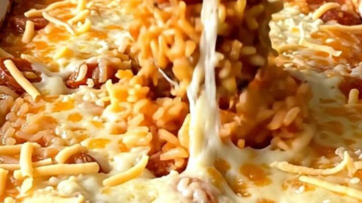 A top-down view of a rectangular baking dish filled with raw rice, crumbled chorizo, tomato broth, and shredded cheese.