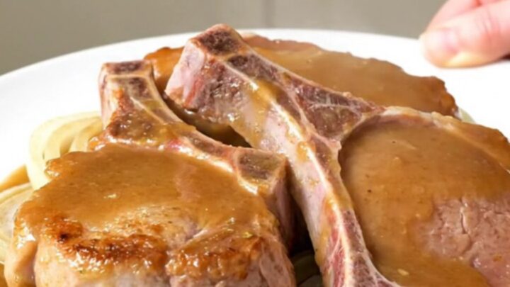 A close-up of tender bone-in pork chops covered in a rich, savory beer and onion gravy on a white plate.