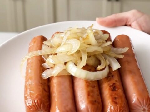 A close-up of tender hot dogs cooking in a slow cooker with sliced onions and beer.