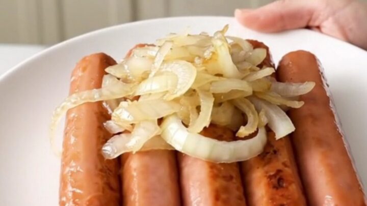 A close-up of tender hot dogs cooking in a slow cooker with sliced onions and beer.