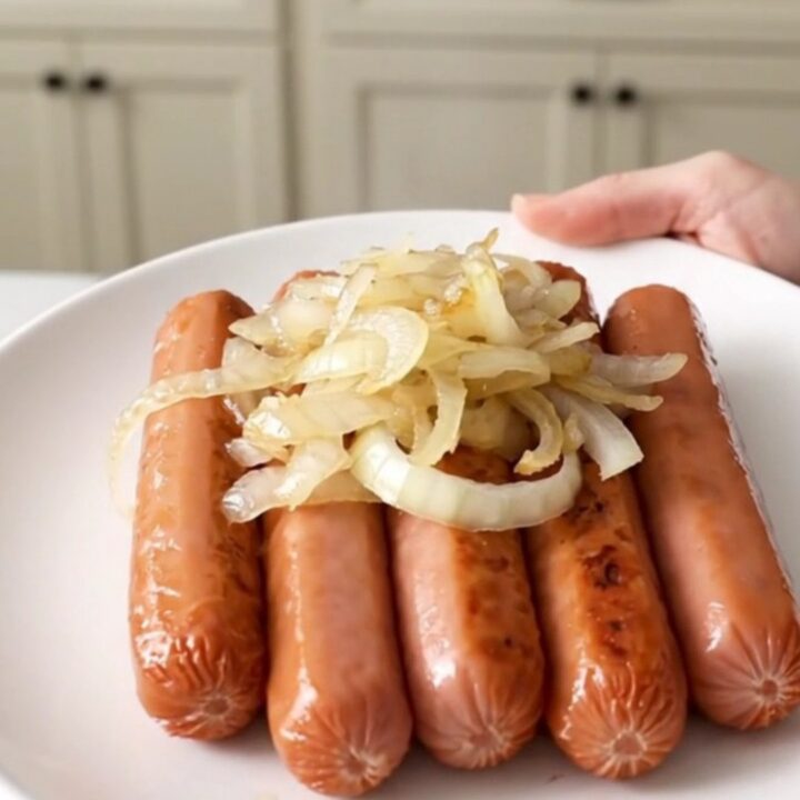 A close-up of tender hot dogs cooking in a slow cooker with sliced onions and beer.