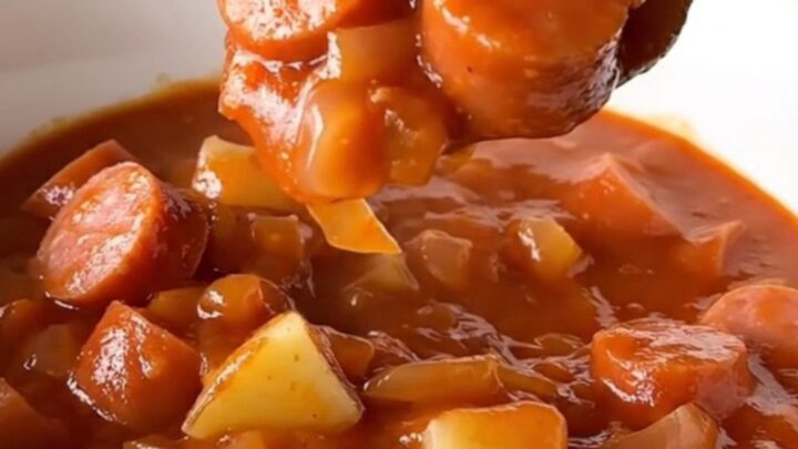 A white shallow bowl filled with a hearty stew of sliced hot dogs and tender potato cubes in a rich, thick red tomato sauce.