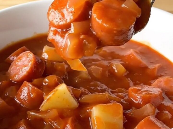 A white shallow bowl filled with a hearty stew of sliced hot dogs and tender potato cubes in a rich, thick red tomato sauce.