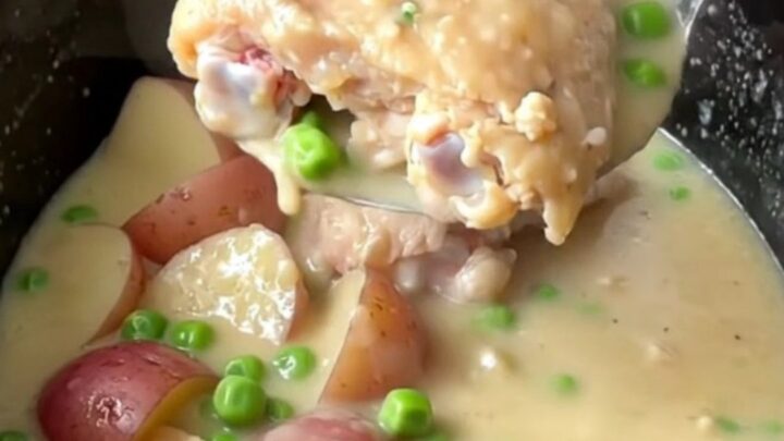 A slow cooker filled with tender chicken thighs, red potatoes, and peas in a rich, creamy white sauce.