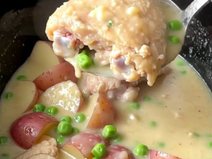A slow cooker filled with tender chicken thighs, red potatoes, and peas in a rich, creamy white sauce.