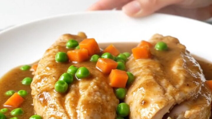 Slow cooker chicken breasts in a rich brown gravy with colorful peas and carrots on a white plate.