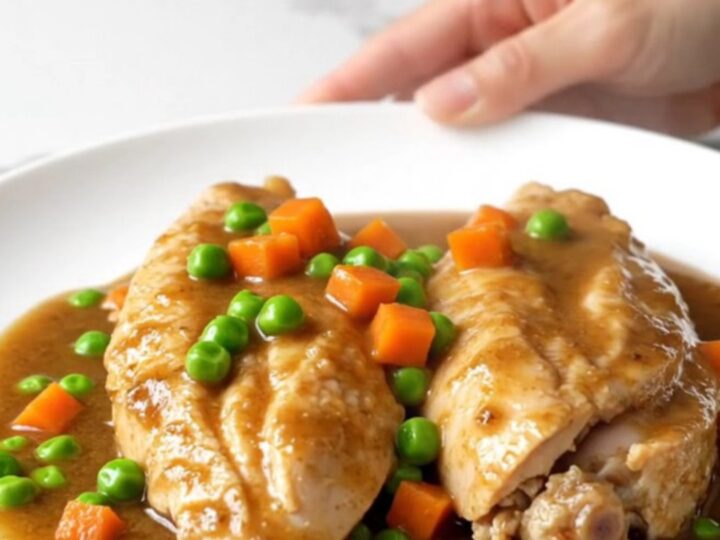 Slow cooker chicken breasts in a rich brown gravy with colorful peas and carrots on a white plate.