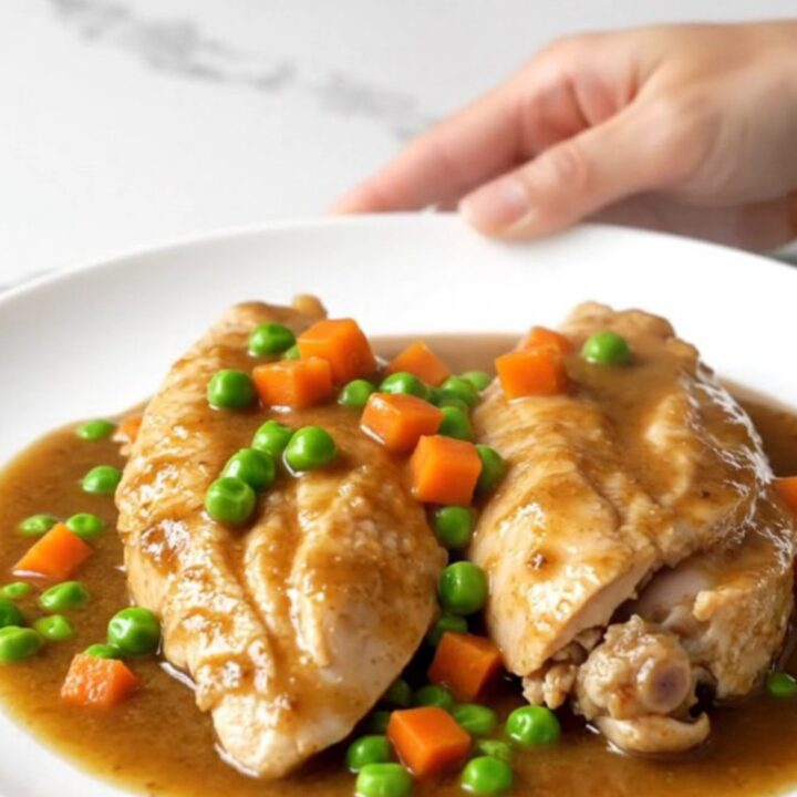 Slow cooker chicken breasts in a rich brown gravy with colorful peas and carrots on a white plate.