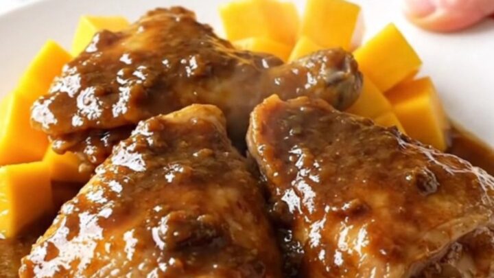 A close-up of tender, braised jerk chicken thighs topped with sweet mango chunks and a glossy sauce.
