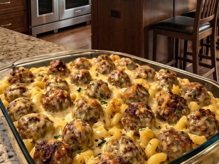 A close-up of a serving spoon lifting a juicy meatball and creamy pasta with a cheese pull from a casserole dish.
