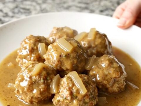 A white plate topped with Swedish chicken meatballs and creamy gravy, with soft-cooked onions, set on a kitchen counter.