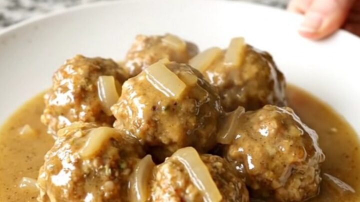 A white plate topped with Swedish chicken meatballs and creamy gravy, with soft-cooked onions, set on a kitchen counter.