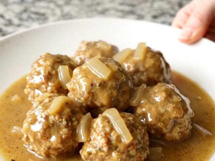 A white plate topped with Swedish chicken meatballs and creamy gravy, with soft-cooked onions, set on a kitchen counter.