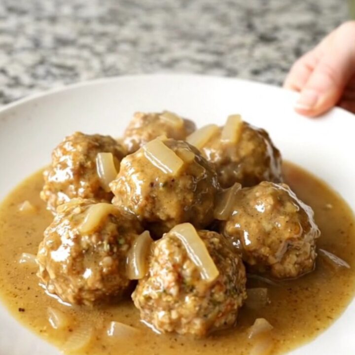 A white plate topped with Swedish chicken meatballs and creamy gravy, with soft-cooked onions, set on a kitchen counter.