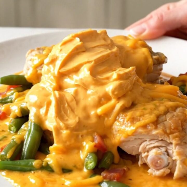 A slow cooker filled with chicken thighs, green beans, bacon, and melted cheddar cheese.