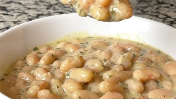 A close-up of creamy garlic parmesan white beans in a white bowl, showing a spoonful being lifted with visible herb flecks.