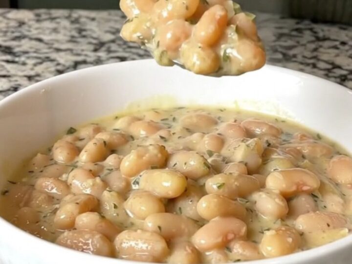 A close-up of creamy garlic parmesan white beans in a white bowl, showing a spoonful being lifted with visible herb flecks.