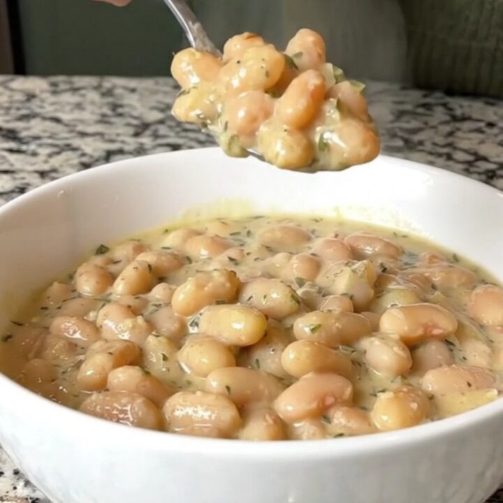 A close-up of creamy garlic parmesan white beans in a white bowl, showing a spoonful being lifted with visible herb flecks.