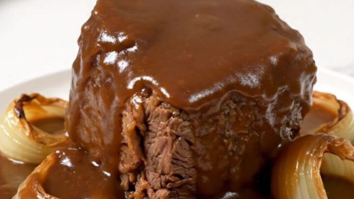 A close-up of a tender beef chuck roast covered in rich brown gravy with soft onions on a white plate.