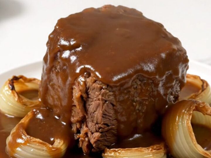 A close-up of a tender beef chuck roast covered in rich brown gravy with soft onions on a white plate.
