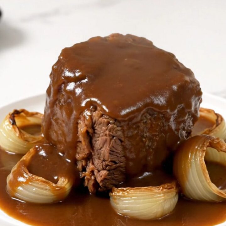 A close-up of a tender beef chuck roast covered in rich brown gravy with soft onions on a white plate.