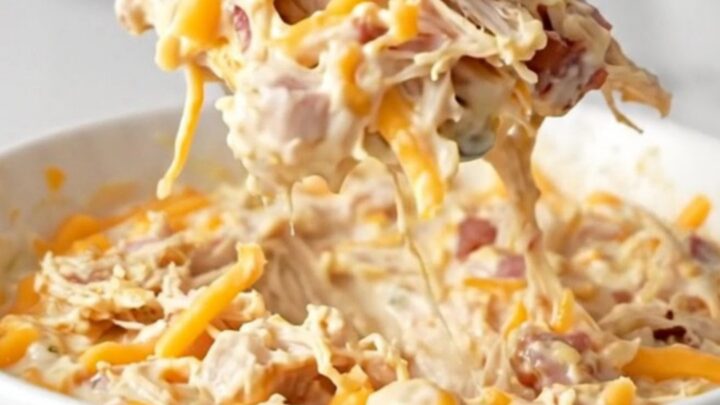 A close-up of a creamy chicken bacon ranch dip in a black slow cooker, featuring melted cheese and visible bacon bits.