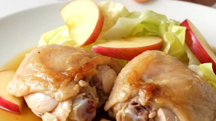 A top-down view of a black slow cooker filled with chicken thighs, cabbage wedges, and apple slices.