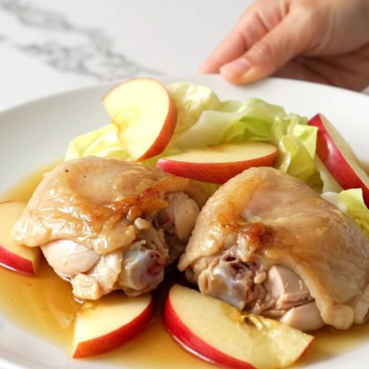 A top-down view of a black slow cooker filled with chicken thighs, cabbage wedges, and apple slices.