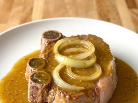 A top-down view of raw bone-in pork shoulder steaks in a white slow cooker insert with onions and sauce.