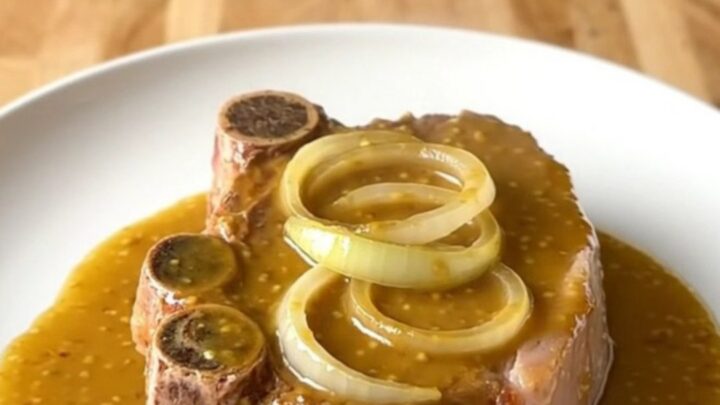 A top-down view of raw bone-in pork shoulder steaks in a white slow cooker insert with onions and sauce.