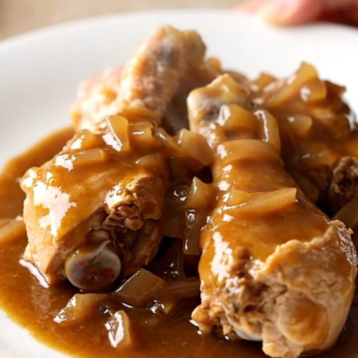 A close-up of tender, slow-cooked chicken drumsticks covered in rich brown gravy, bell peppers, and onions.