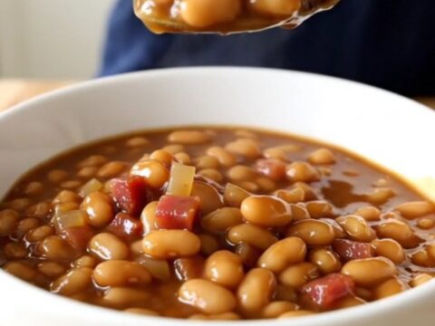 A white ceramic slow cooker filled with slow-cooked Boston baked beans, showing a rich, dark, molasses-based sauce.