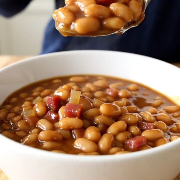 A white ceramic slow cooker filled with slow-cooked Boston baked beans, showing a rich, dark, molasses-based sauce.