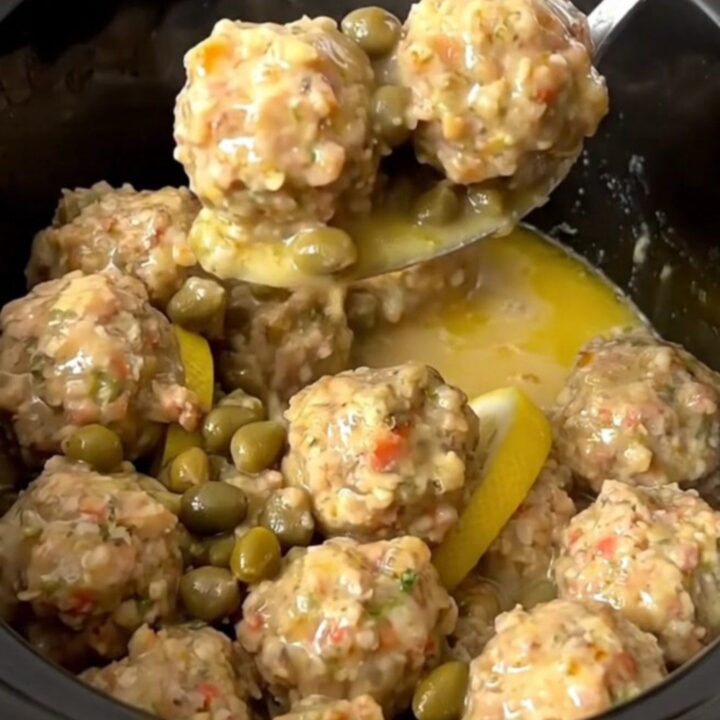 A close-up of tender chicken meatballs in a glossy lemon and caper butter sauce in a slow cooker.