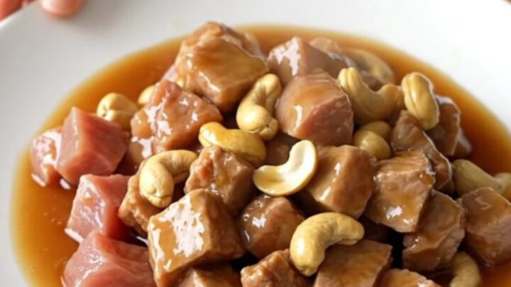A white slow cooker insert filled with cubes of tender pork loin and whole roasted cashews in a glossy honey soy sauce.