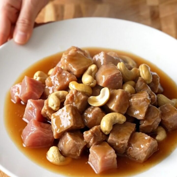 A white slow cooker insert filled with cubes of tender pork loin and whole roasted cashews in a glossy honey soy sauce.