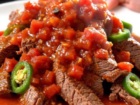 A close-up of tender, slow-cooked round steak strips in a rich tomato sauce with golden potatoes and jalapeños.
