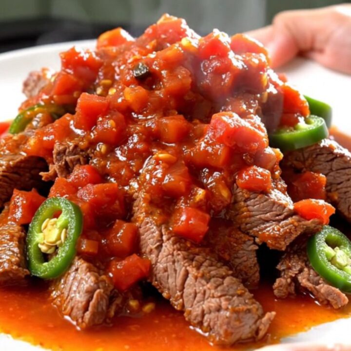 A close-up of tender, slow-cooked round steak strips in a rich tomato sauce with golden potatoes and jalapeños.