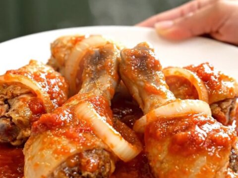A close-up of tender chicken drumsticks braised in a thick, red tomato sauce with visible onion slices in a slow cooker.