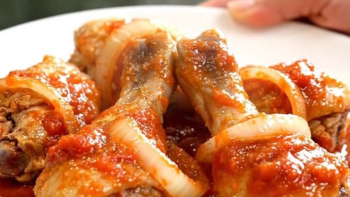 A close-up of tender chicken drumsticks braised in a thick, red tomato sauce with visible onion slices in a slow cooker.