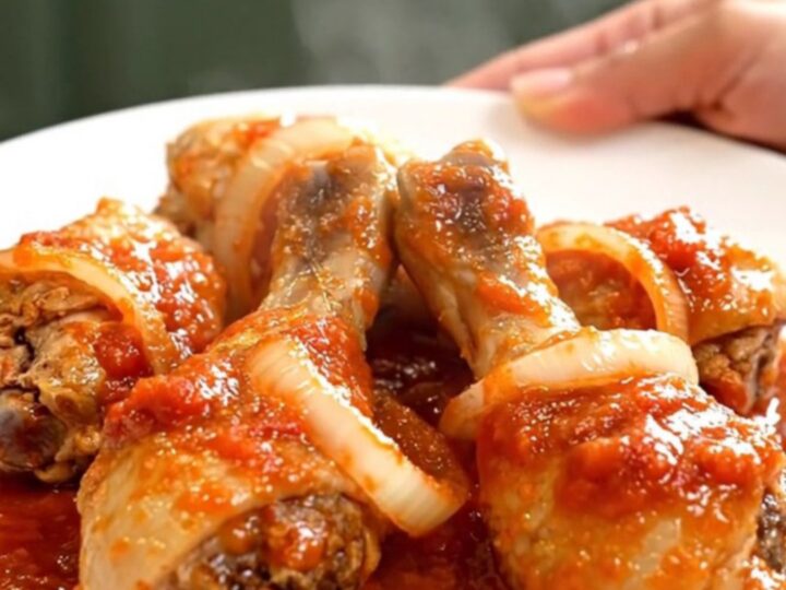 A close-up of tender chicken drumsticks braised in a thick, red tomato sauce with visible onion slices in a slow cooker.