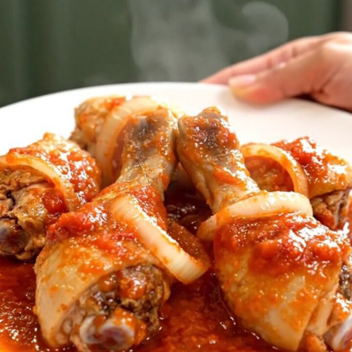 A close-up of tender chicken drumsticks braised in a thick, red tomato sauce with visible onion slices in a slow cooker.