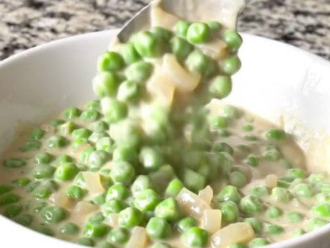A shallow white bowl filled with creamy peas and pearl onions in a thick white sauce.
