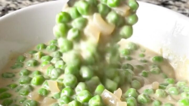 A shallow white bowl filled with creamy peas and pearl onions in a thick white sauce.