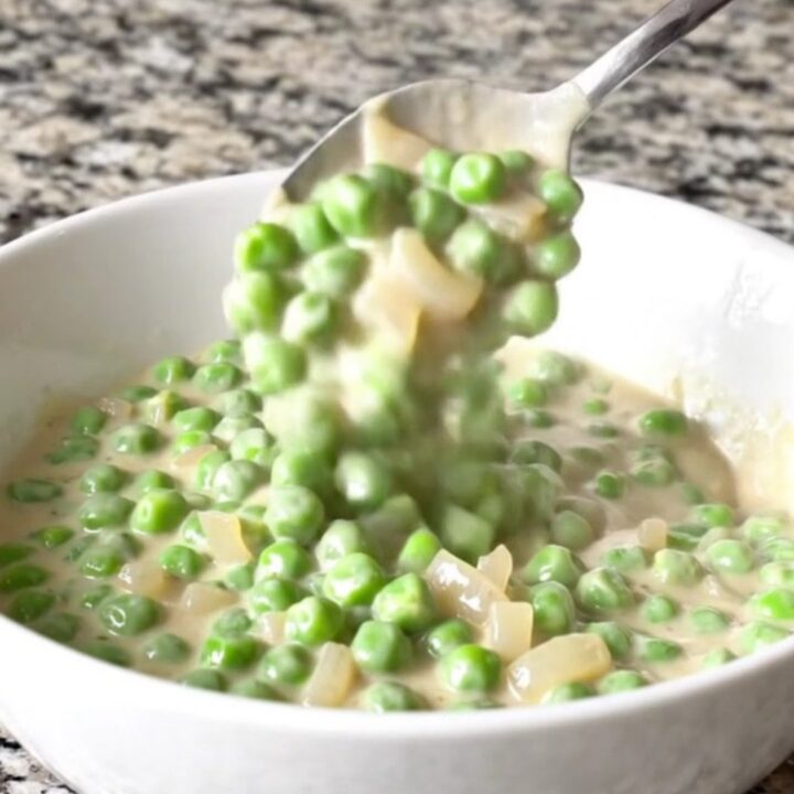 A shallow white bowl filled with creamy peas and pearl onions in a thick white sauce.