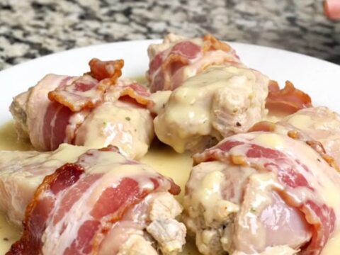 Bacon-wrapped chicken breasts resting in a creamy white sauce inside a matte black slow cooker.