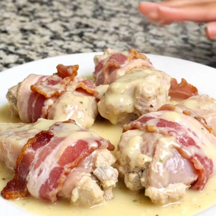 Bacon-wrapped chicken breasts resting in a creamy white sauce inside a matte black slow cooker.